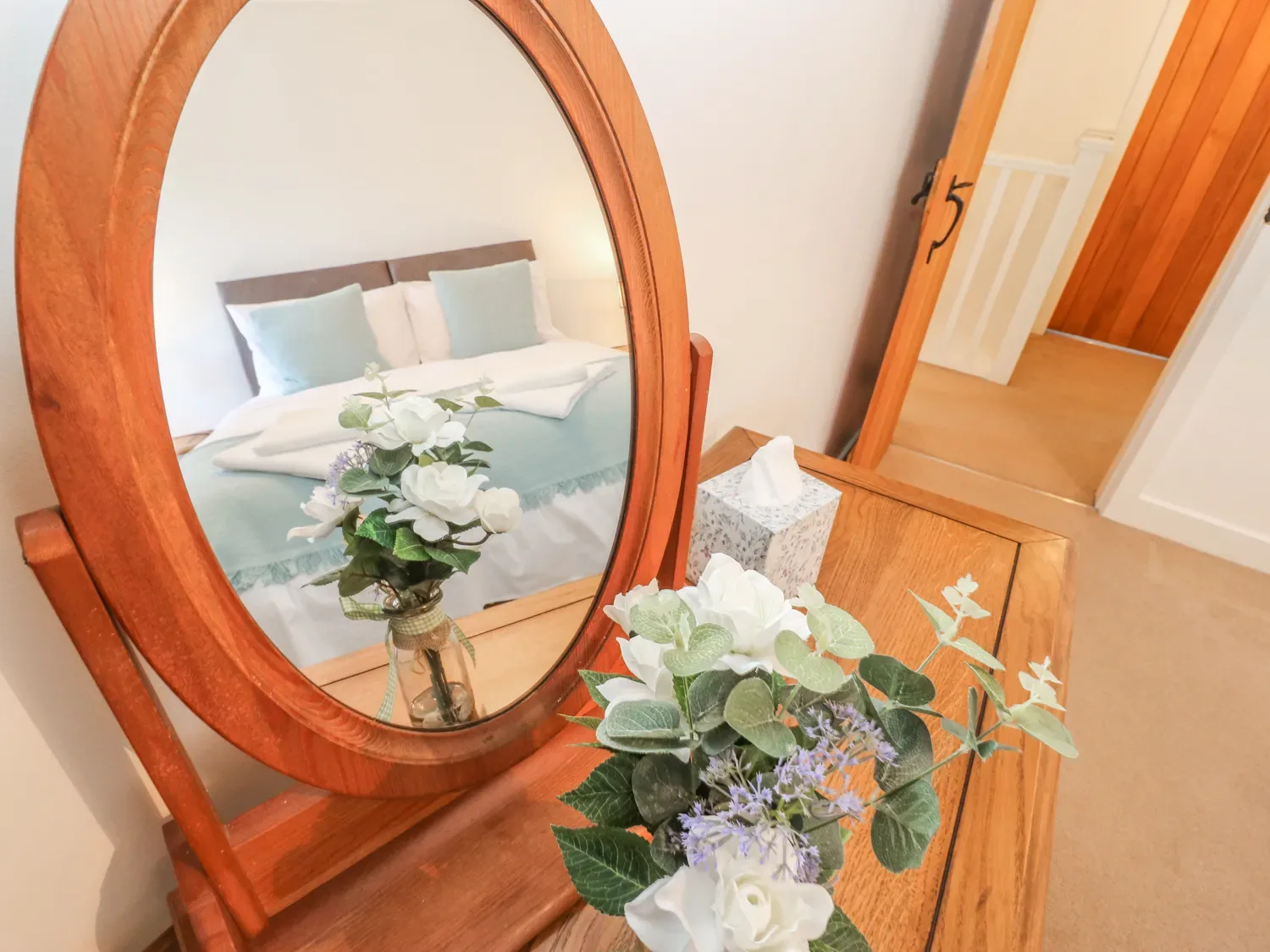 A wooden dressing table with a round mirror flowers on the table and a reflection of a bed in a bedroom at The Cottage Beeston Hall in Ripponden