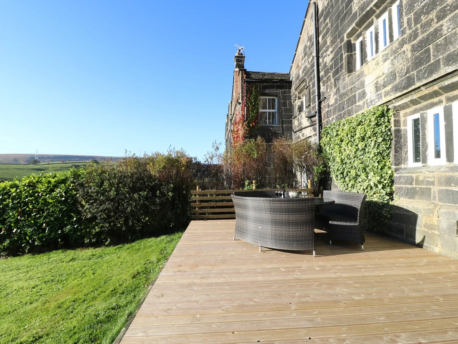 A wooden deck with outdoor seating beside a stone building and leafy hedges.