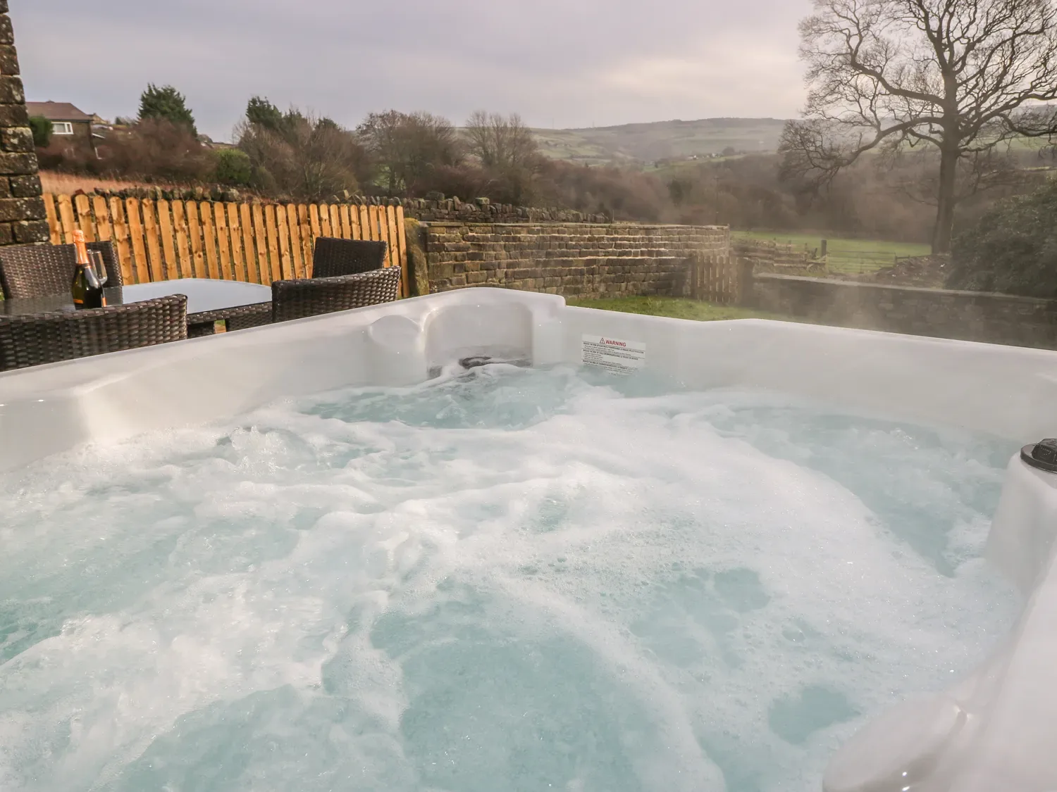 An outdoor hot tub with bubbling water overlooks a stone wall and rolling countryside.