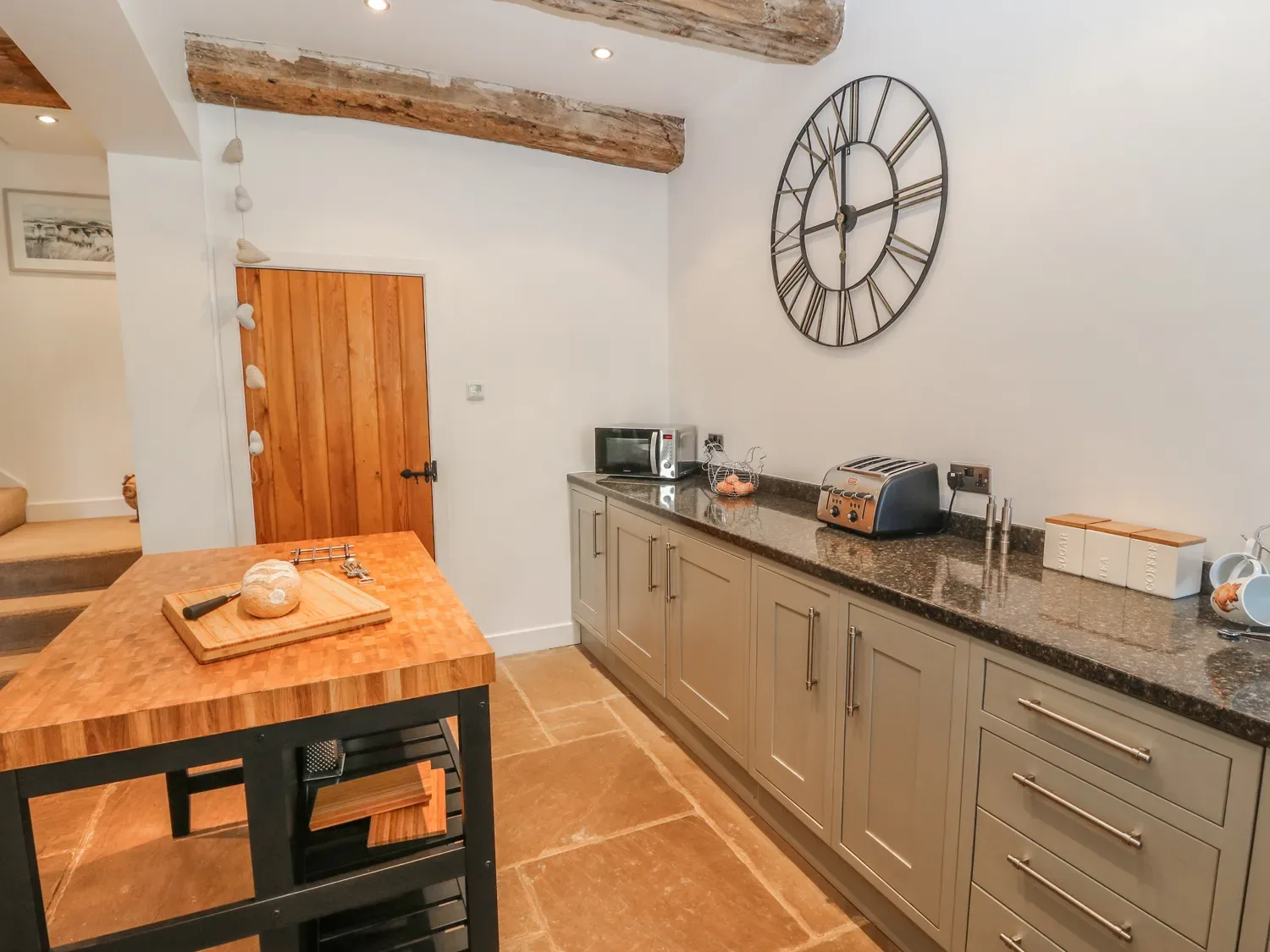 A kitchen with a wooden island table a loaf of bread on a cutting board a microwave toaster and large clock on the wall at The Cottage Beeston Hall Ripponden