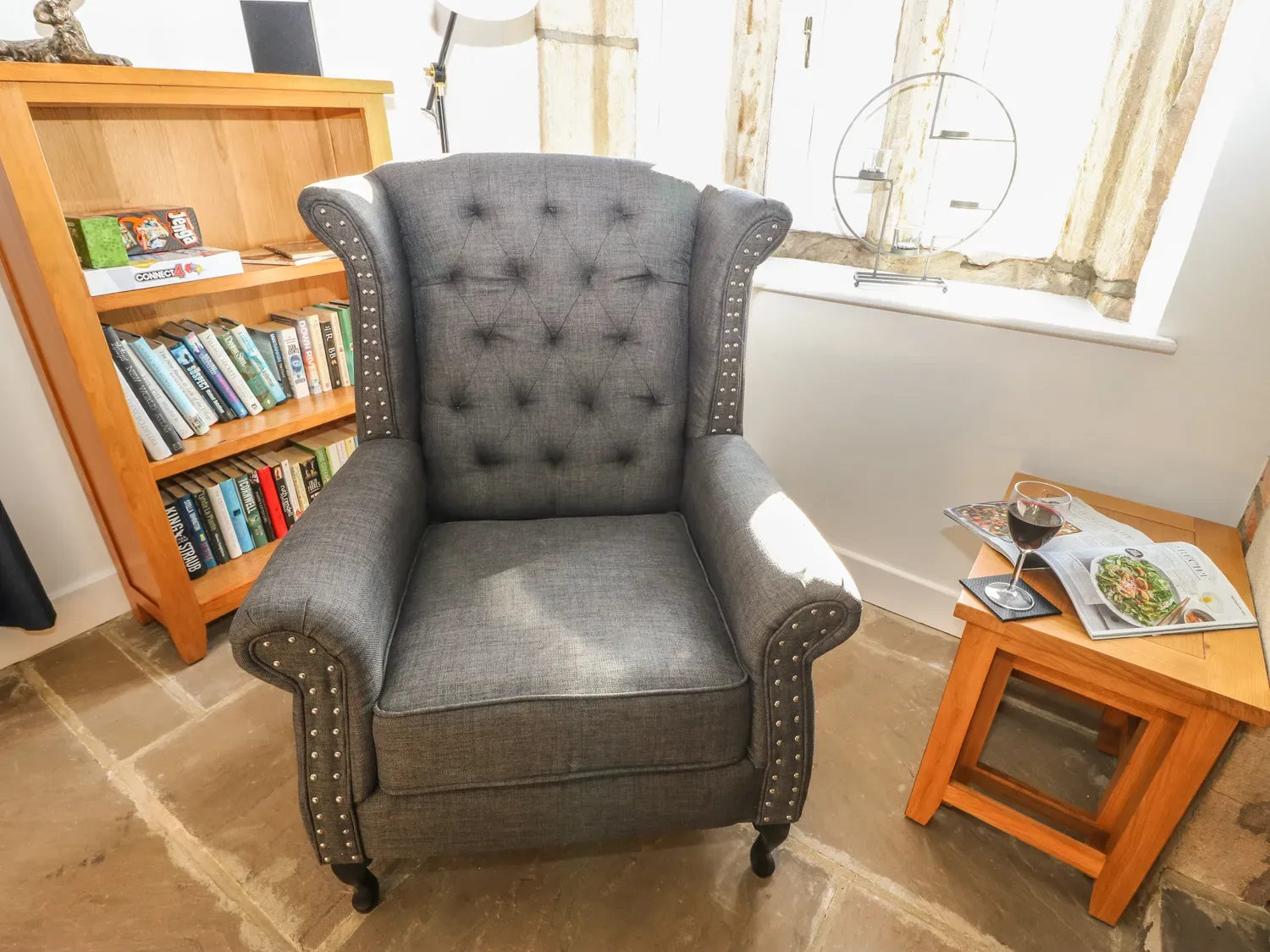 A gray armchair next to a wooden side table with a glass of red wine and magazines near a window and a wooden bookshelf with books at The Cottage Beeston Hall in Ripponden
