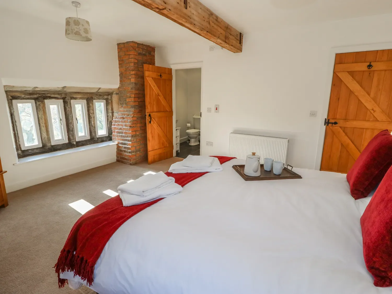 A bedroom with a bed covered in white and red linens two wooden doors a window with multiple small panes and a bathroom visible in the background at The Cottage Beeston Hall in Ripponden