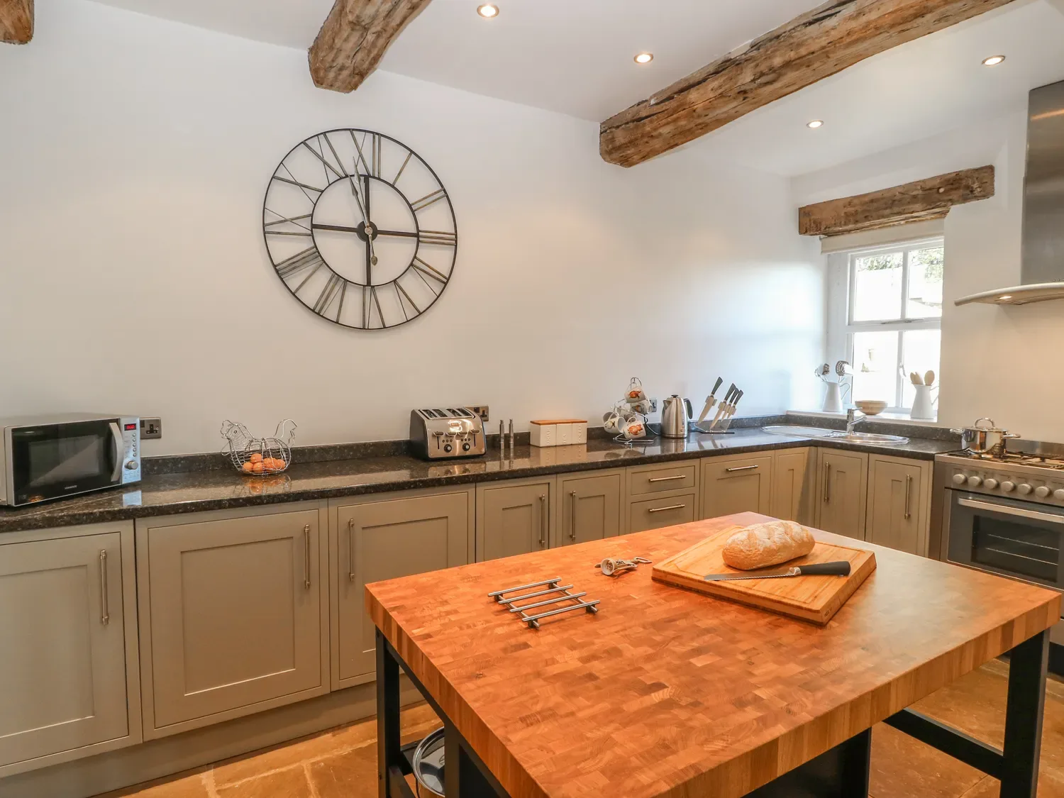 A kitchen with wooden beams on the ceiling a large clock on the wall cabinets a microwave toaster a kettle and a wooden island with bread and knife at The Cottage, Beeston Hall in Ripponden