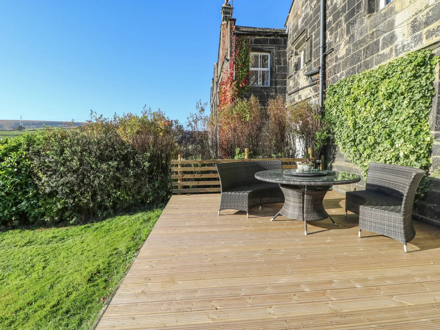 A wooden outdoor deck with a round glass table and two wicker chairs next to a stone building with ivy at The Cottage Beeston Hall in Ripponden