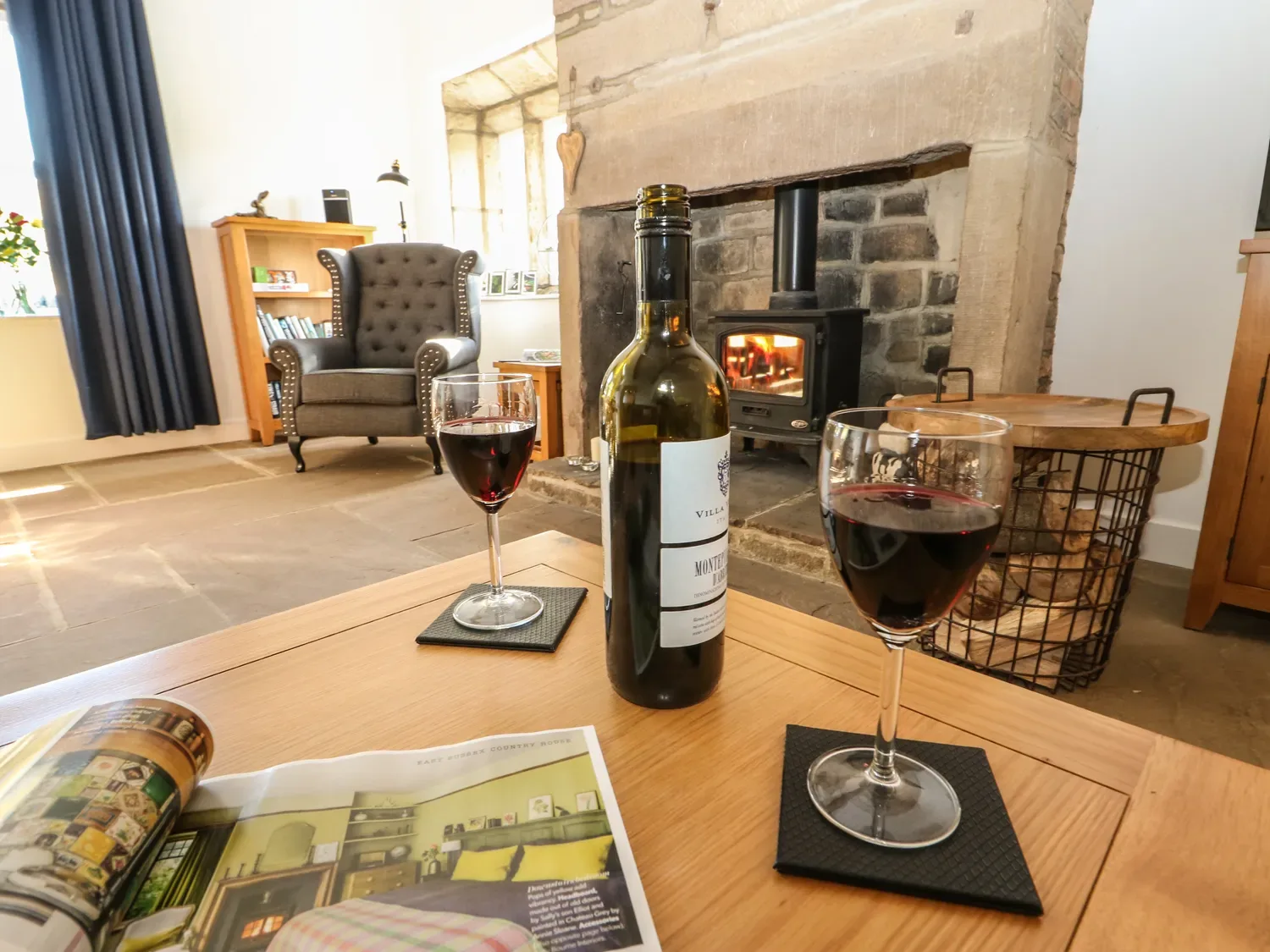 A living room with wine on a table and a wood-burning stove beside the fireplace.
