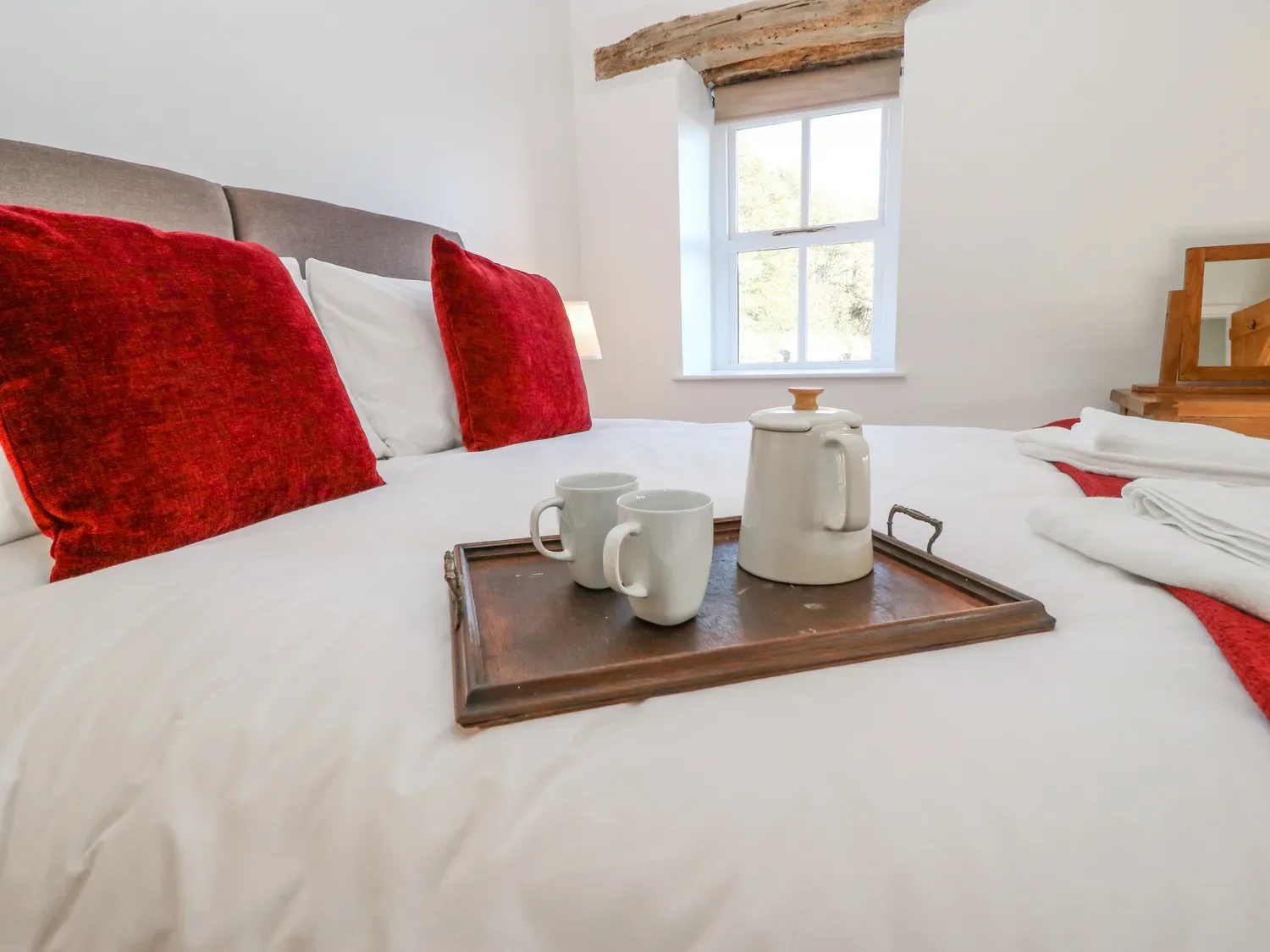 A bedroom with a bed with white linens and red cushions with a tray holding a teapot and two cups a window and a wooden dresser at The Cottage Beeston Hall Ripponden
