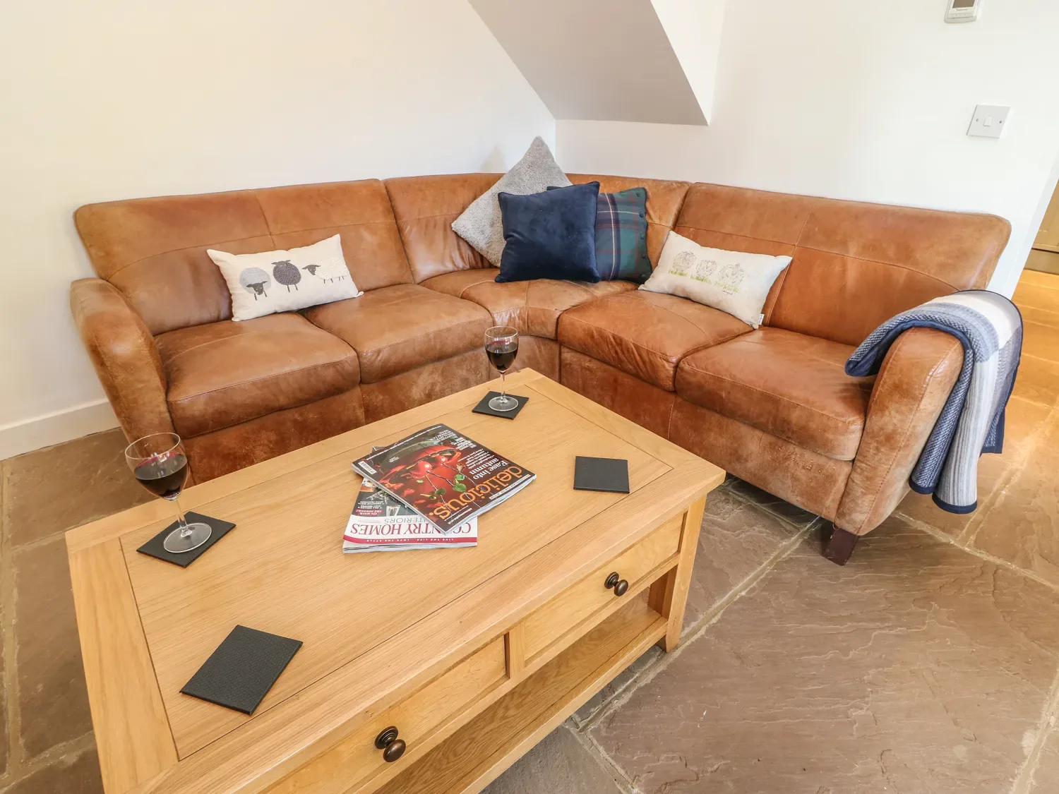 A corner sofa with cushions sits behind a wooden coffee table holding magazines and wine glasses.
