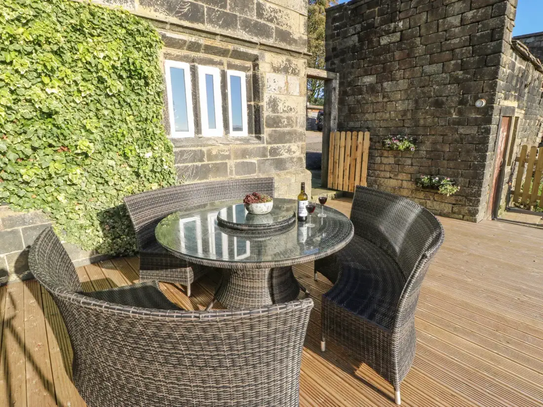 An outdoor patio with a round glass table and curved wicker chairs on a wooden deck with stone buildings and ivy at The Cottage Beeston Hall in Ripponden