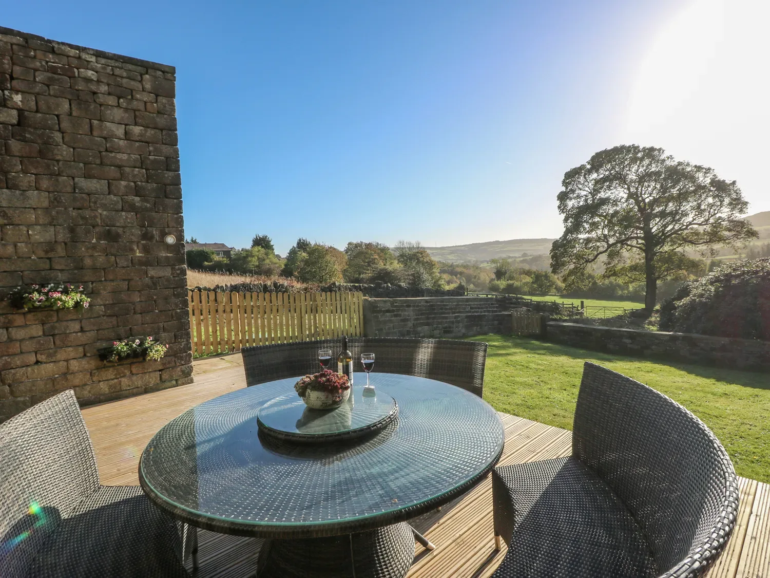 An outdoor patio with a round glass table and wicker chairs overlooking a grassy yard and scenic landscape at The Cottage Beeston Hall in Ripponden