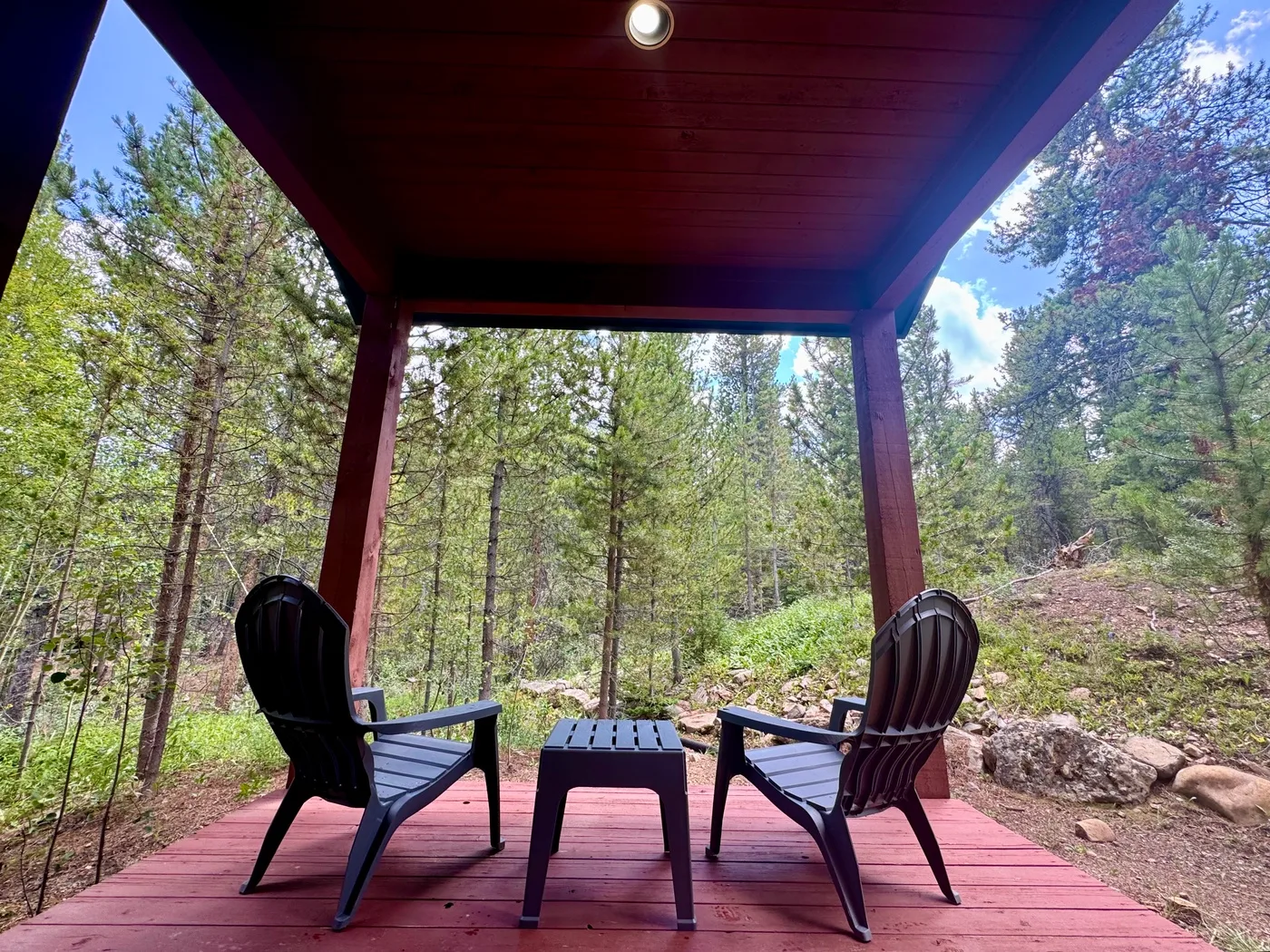 A covered wooden deck with two chairs and a small table overlooks a wooded landscape.