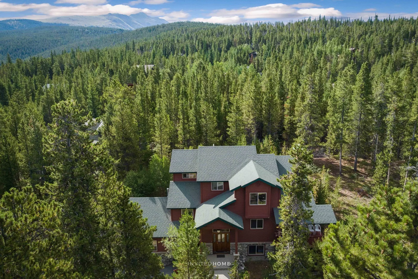 A red house sits among dense evergreen trees, with forested hills in the distance.