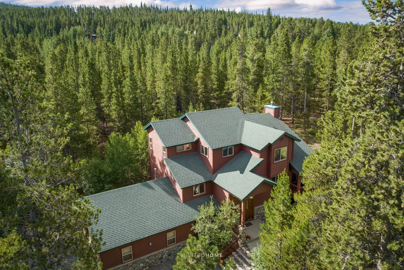A red multi-level house with green roofs sits among dense evergreen trees.