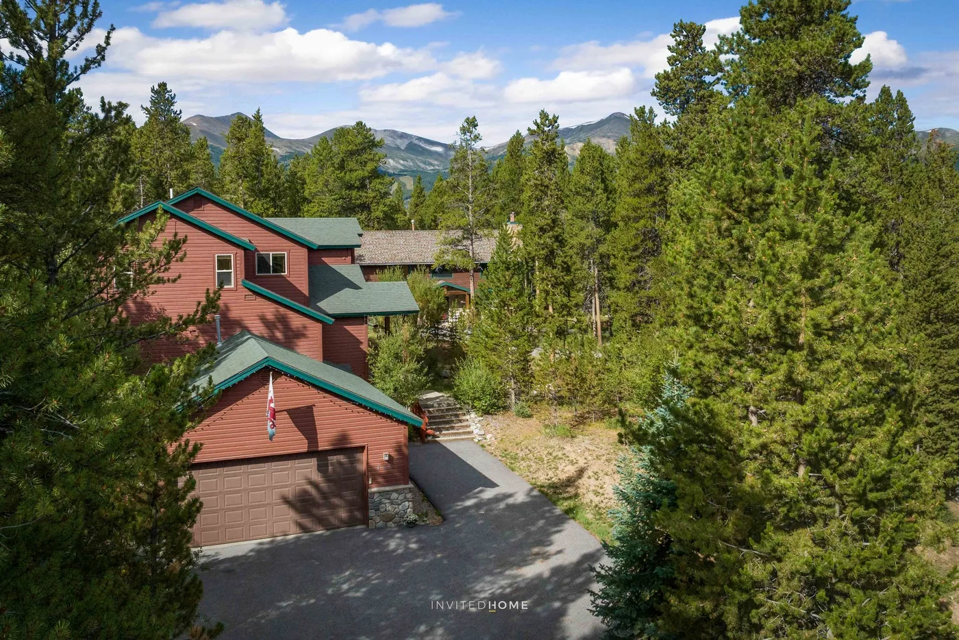 A multi-level house with green roofs stands among pine trees, with mountains in the distance.