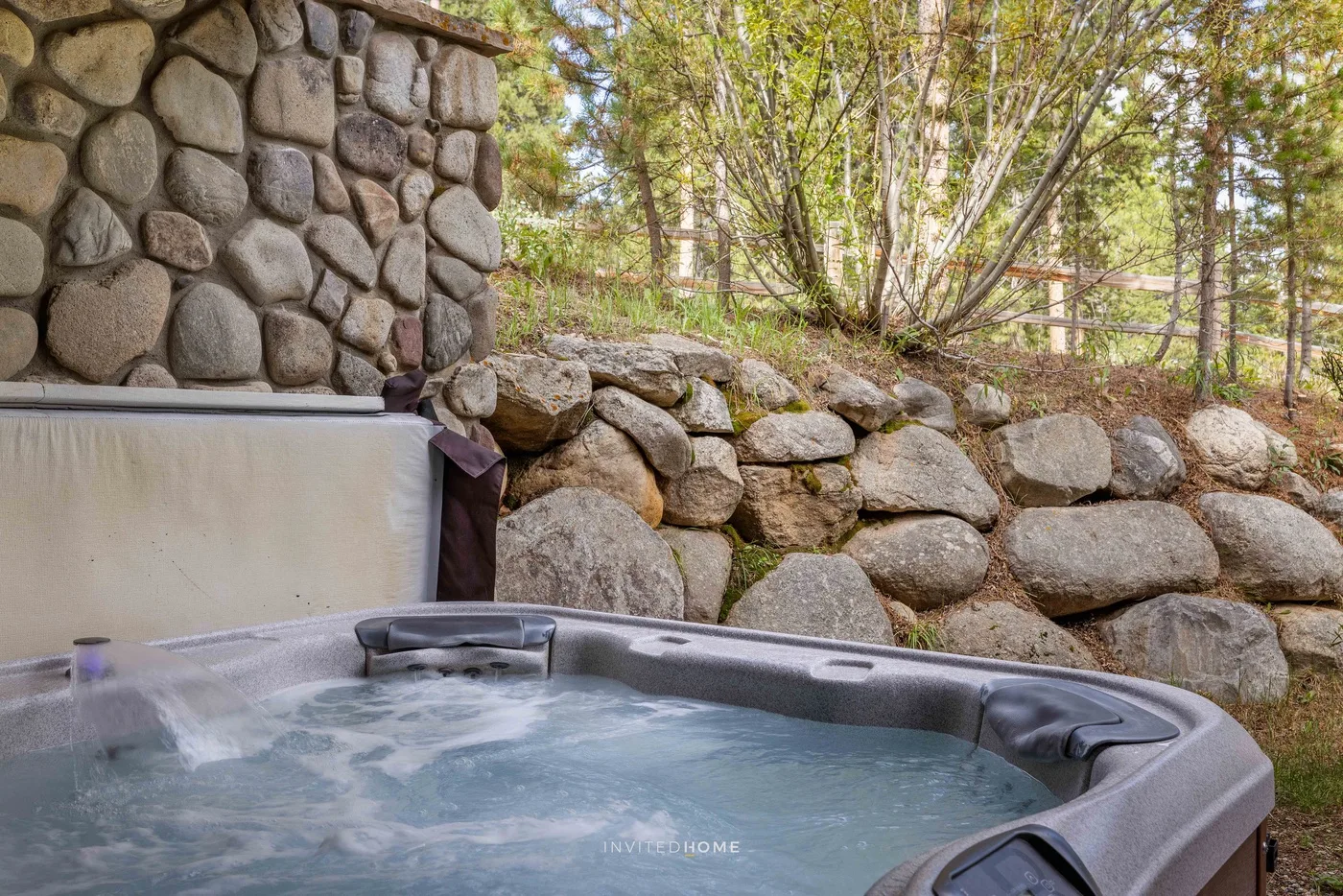 An outdoor hot tub sits beside a stone wall, with trees in the background.
