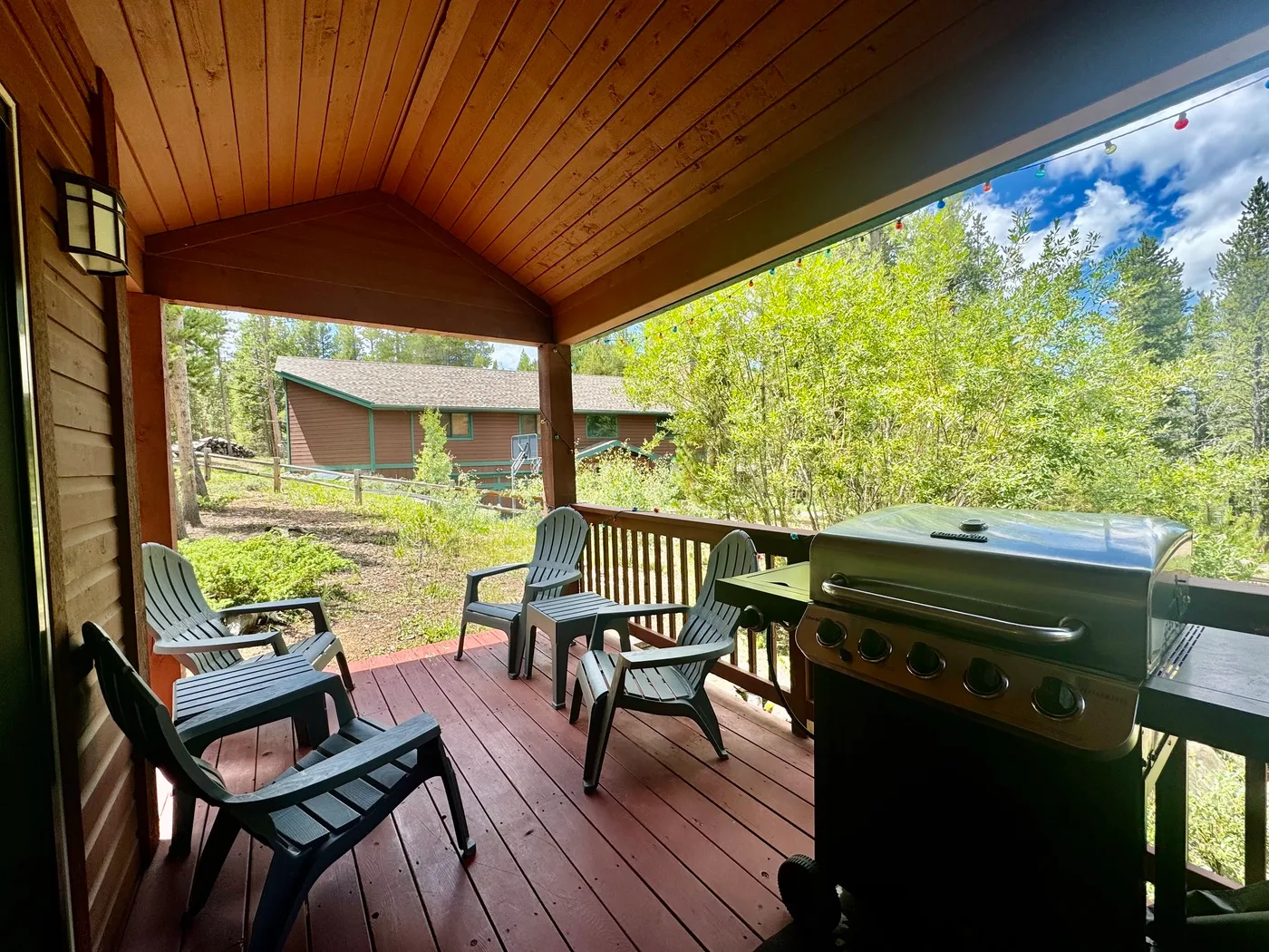 A covered wooden deck has outdoor seating and a barbecue grill overlooking trees.