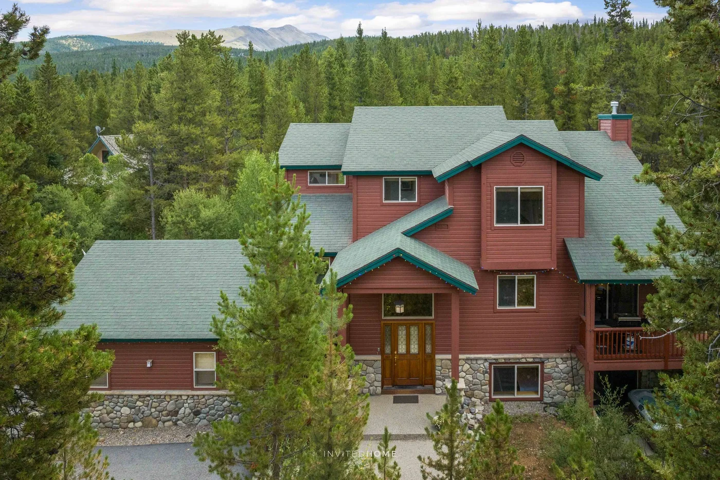 A large red house with green roofs stands among trees against a mountain backdrop.