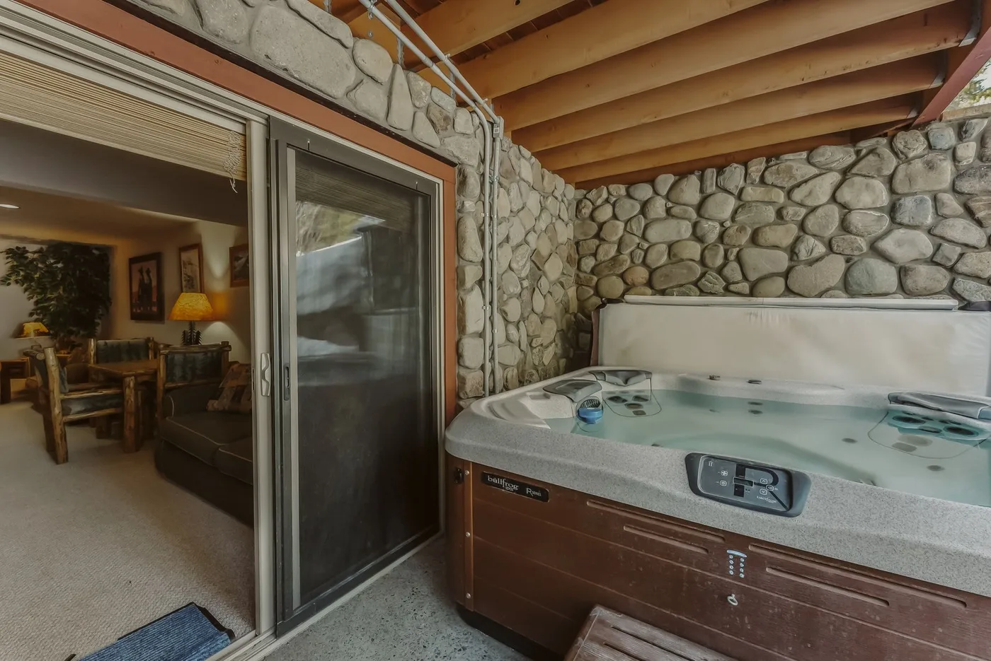 A covered patio with a hot tub beside a sliding glass door and stone walls.