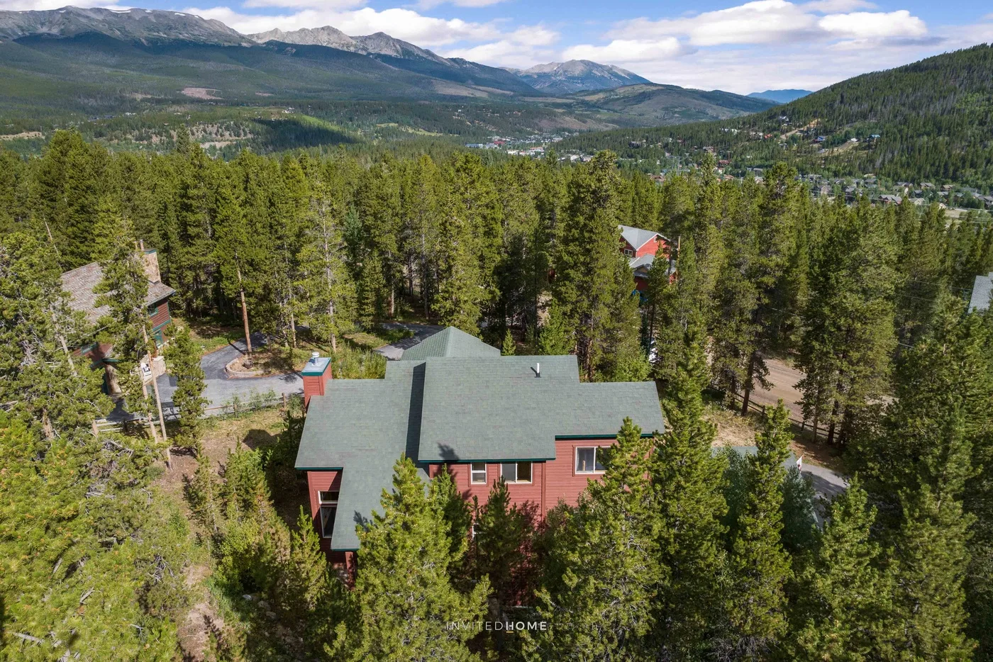 An aerial view of a red house among pine trees, with mountains in the distance.
