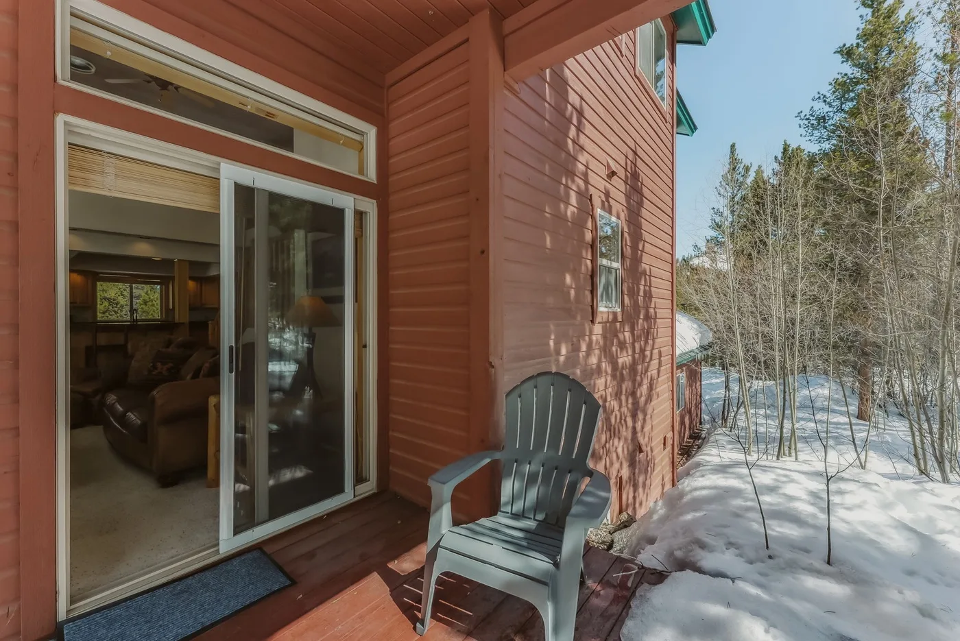 A snowy outdoor deck beside a red-sided cabin, with a chair near the sliding glass door.