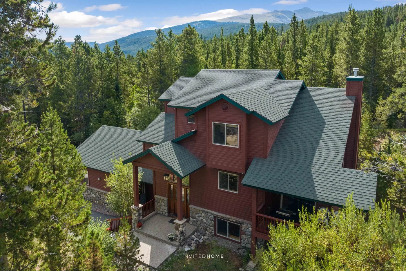 A large red house with multiple gabled roofs sits among trees, with mountains in the distance.