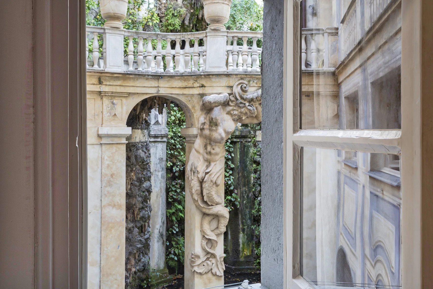 Una ventana abierta da a un patio de piedra con una figura esculpida y vegetación.