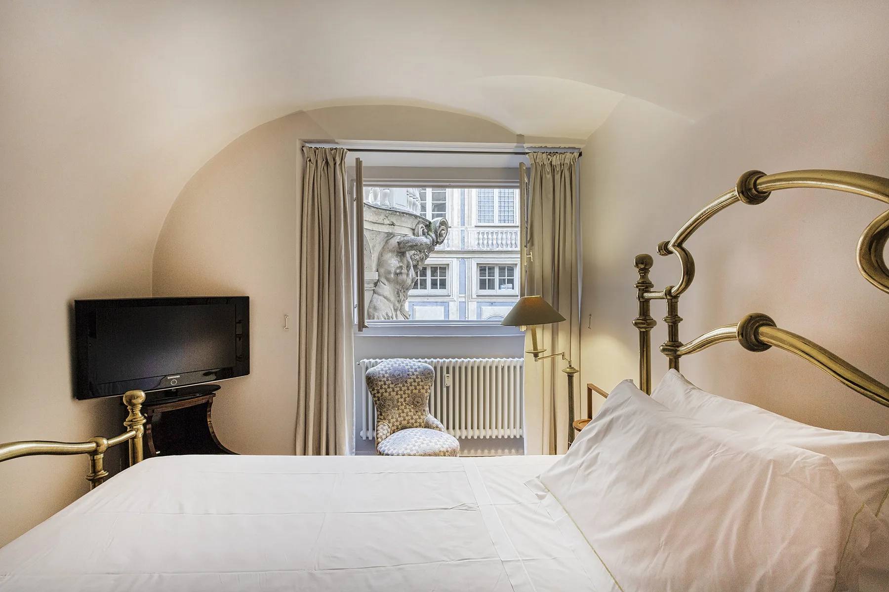 Un dormitorio con una cama tendida, cabecero de latón y una ventana con edificios cercanos a la vista.