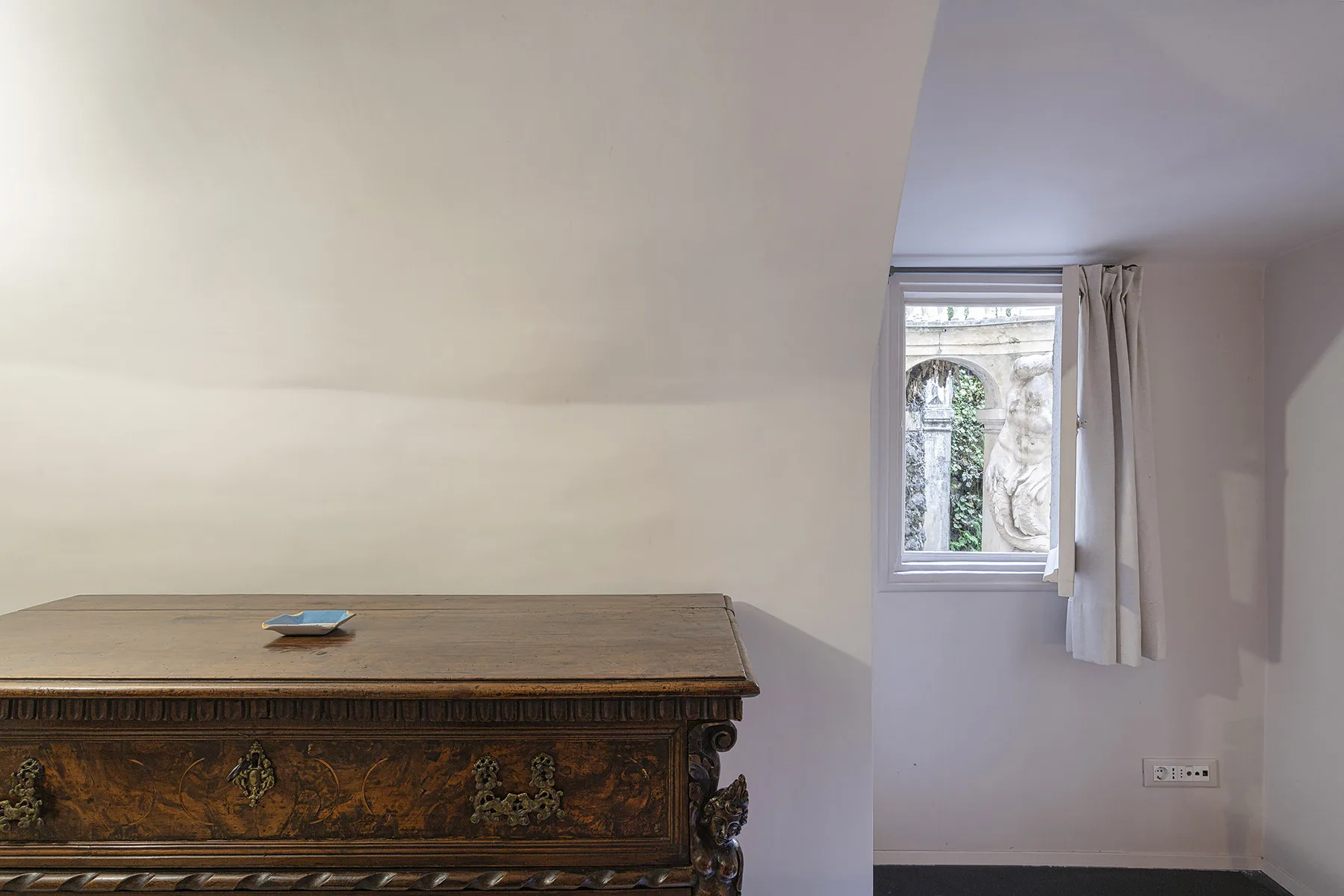 Un rincón de dormitorio con un baúl de madera ornamentado y una ventana con cortinas.