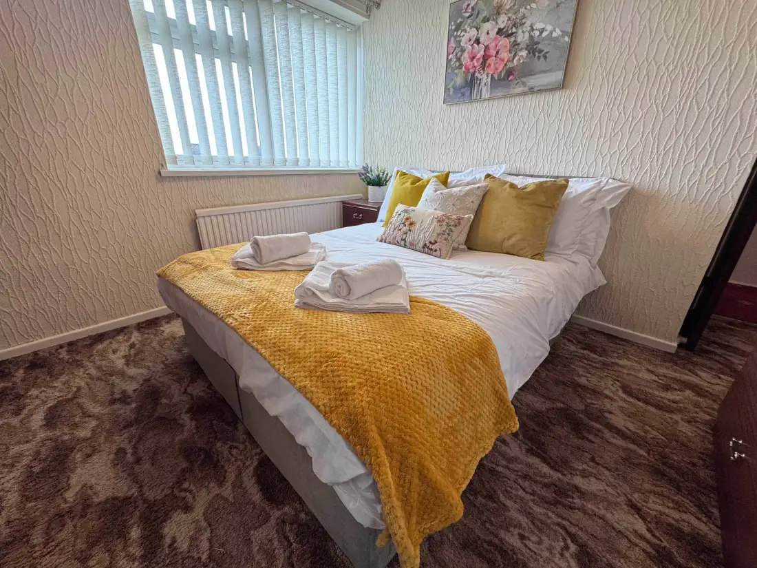A bedroom with a neatly made bed, mustard-yellow throw, and window blinds.