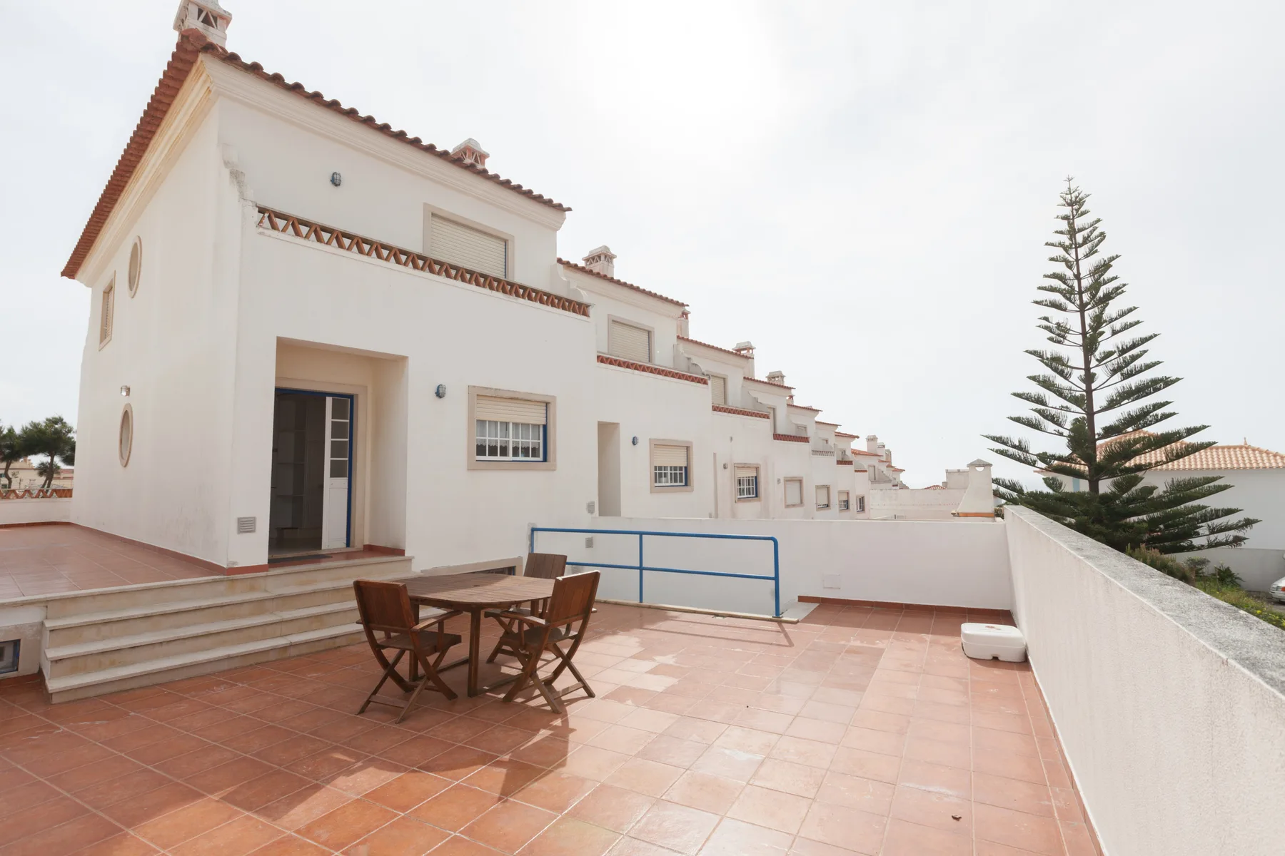 A tiled outdoor terrace with a wooden dining table beside a white building.