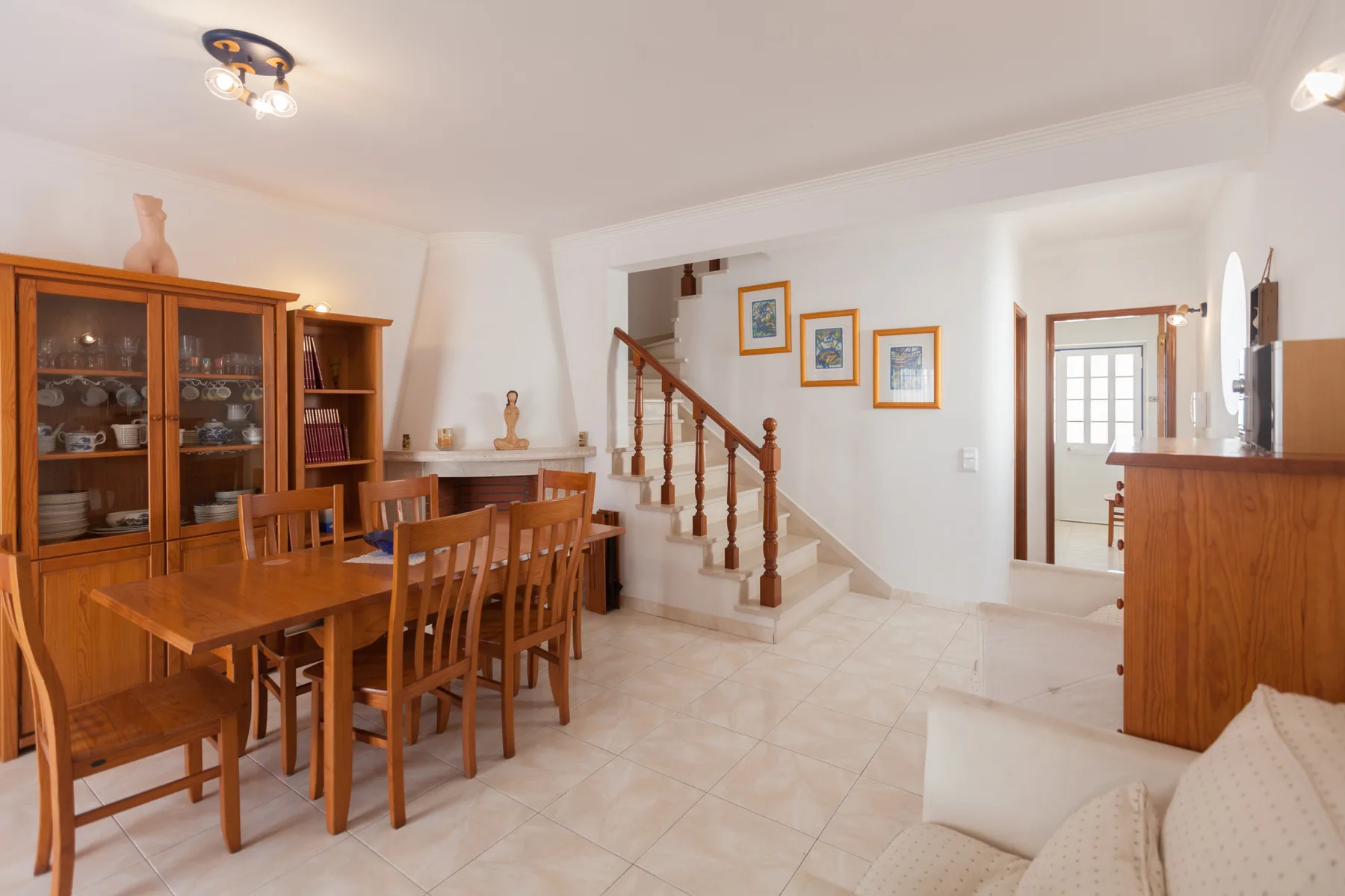 A dining area with wooden seating beside a staircase and a bright adjoining room.