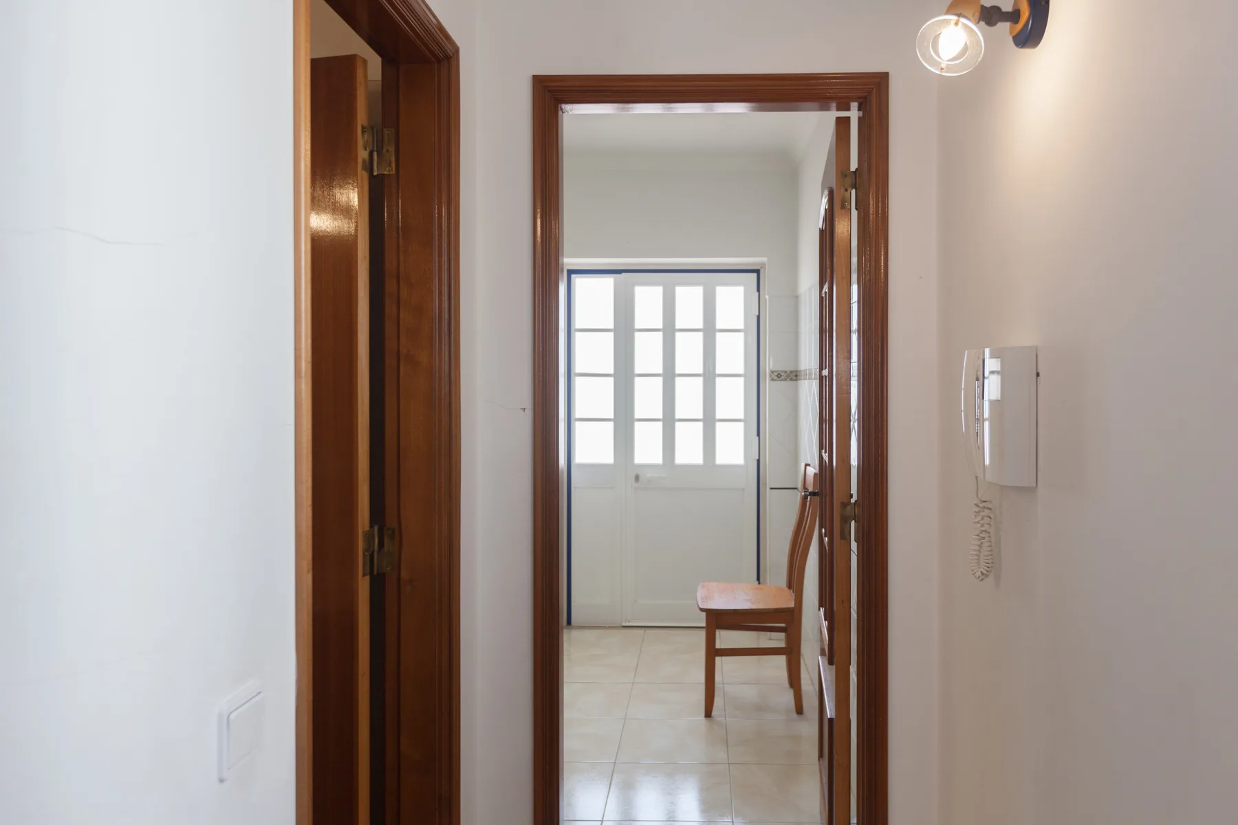 Un pasillo interior da a una habitación luminosa con una silla junto a la ventana.
