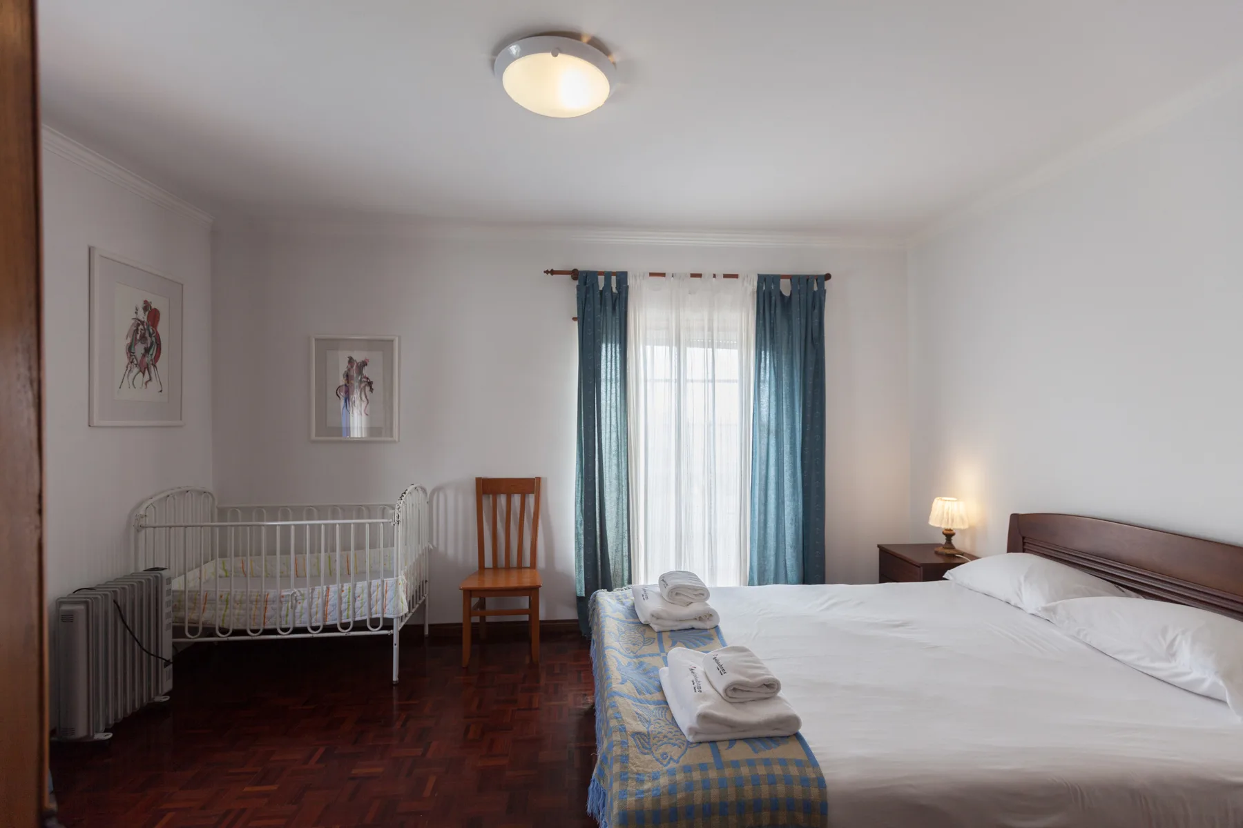 A bedroom with a large bed, a crib, and a window with blue curtains.