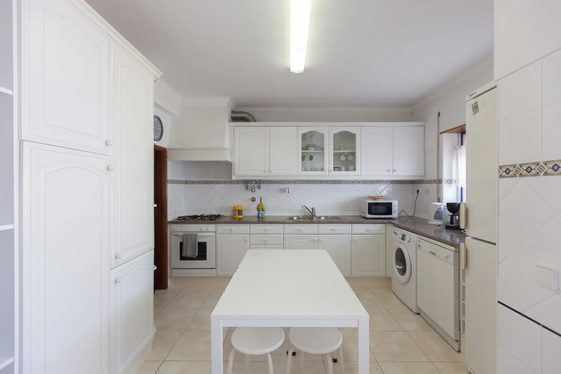 A bright kitchen with white cabinets, a central table, and appliances.