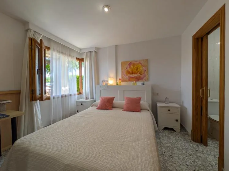 Un dormitorio con una cama tendida, dos almohadas y ventanas altas con cortinas translúcidas.