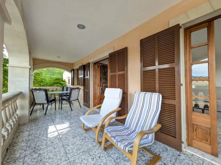 A covered veranda with cushioned seating and wooden shutters along the wall.