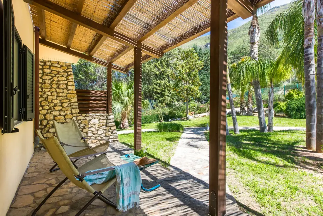 A shaded patio with reclining seating overlooks a garden path and palm trees.
