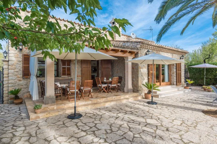 A stone house opens onto a paved patio with outdoor seating and umbrellas.