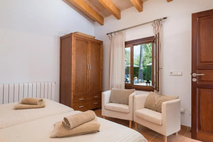 A bedroom with two beds, wooden storage, and seating beside a window.