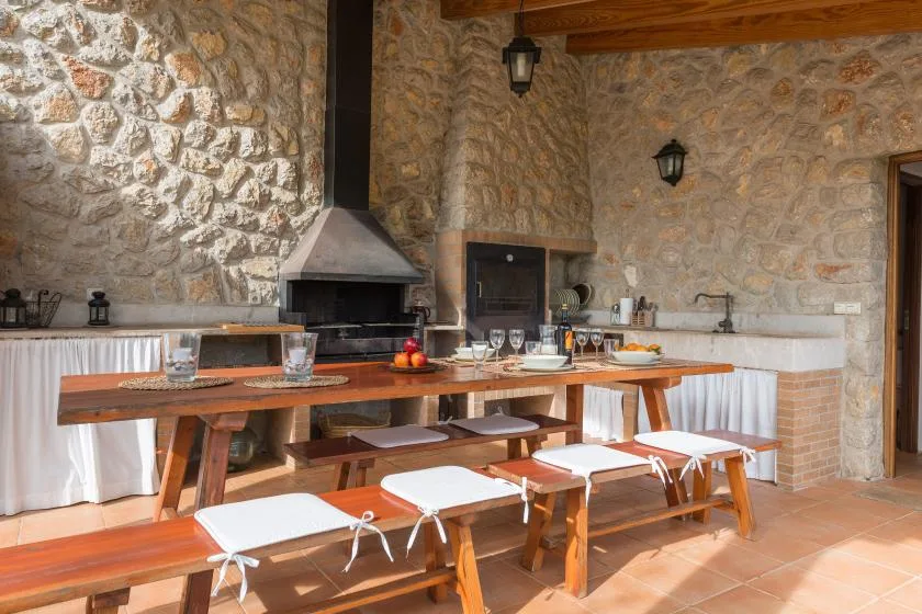 A stone-walled dining area has long wooden tables and benches beside an outdoor kitchen.