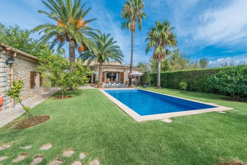 A garden pool beside a stone house, with palm trees and green lawn.