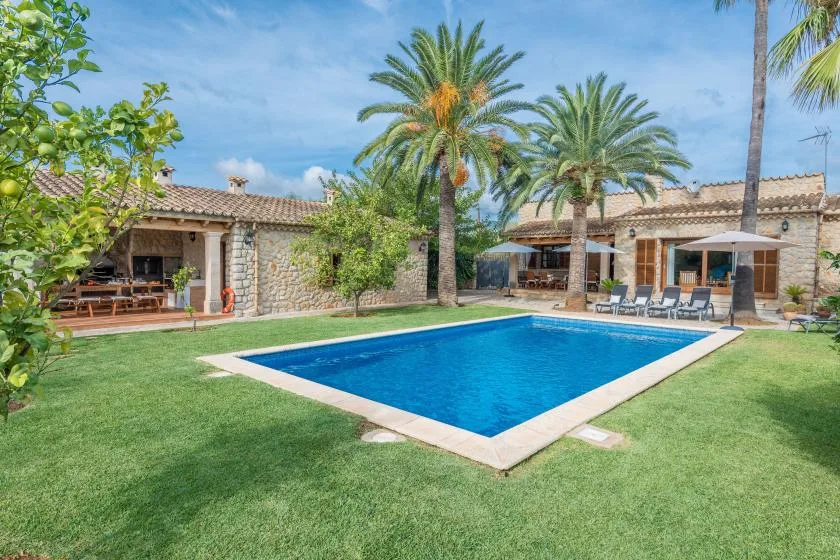 A swimming pool sits in a grassy garden beside a stone house and palm trees.