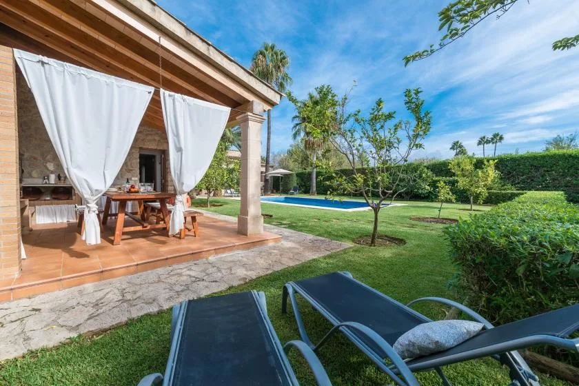A covered patio with dining furniture overlooks a green garden and swimming pool.