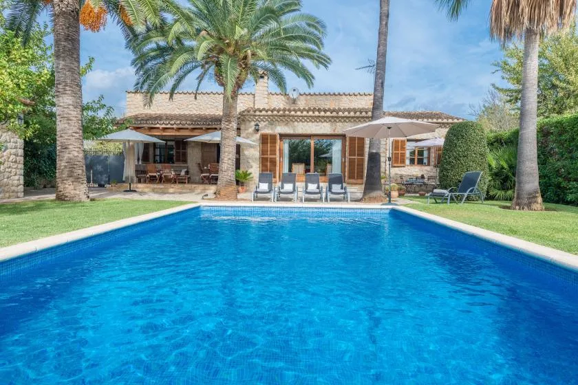 A swimming pool in front of a house, with patio seating and palm trees.