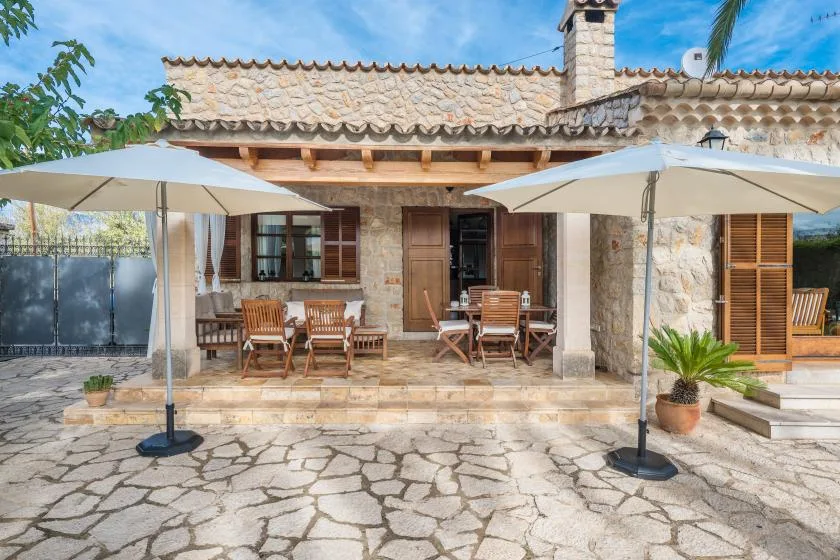 A stone house opens onto a sunny patio with outdoor seating and umbrellas.