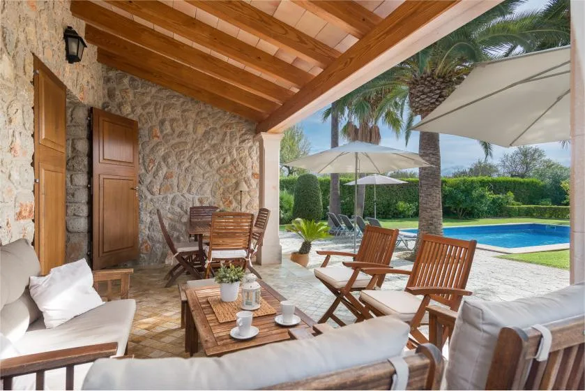 A covered stone patio with seating overlooks a swimming pool and garden.