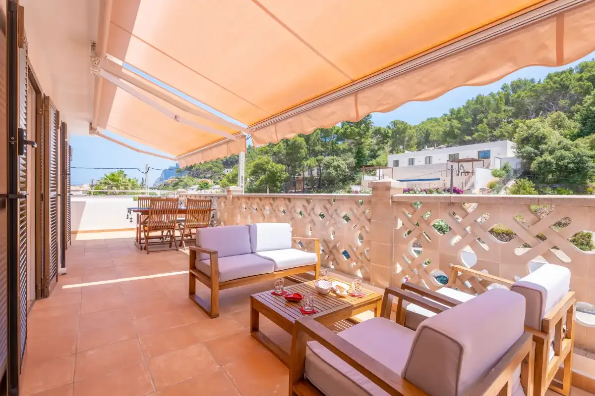 Una terraza exterior cubierta con asientos y una mesa de comedor da a árboles y edificios cercanos.