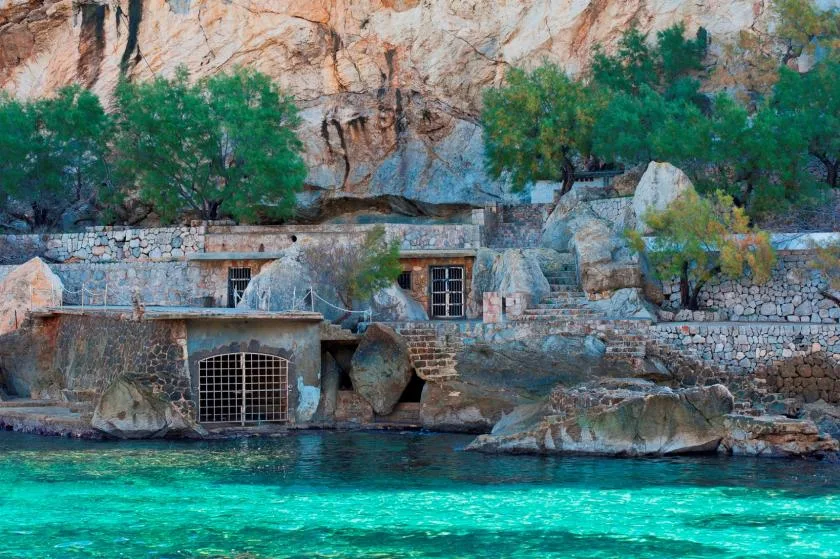 Un edificio de piedra se encuentra junto a aguas turquesas bajo un acantilado rocoso.