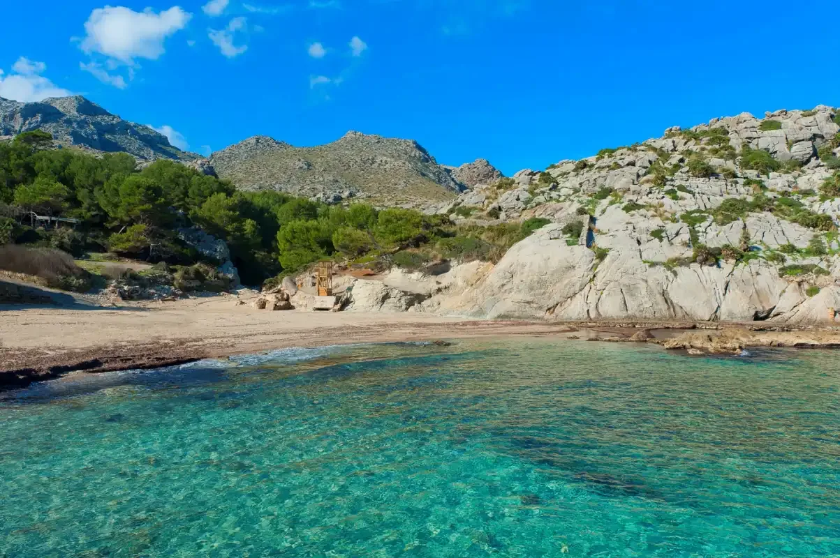 Una pequeña cala con agua turquesa clara, una orilla arenosa y colinas rocosas.