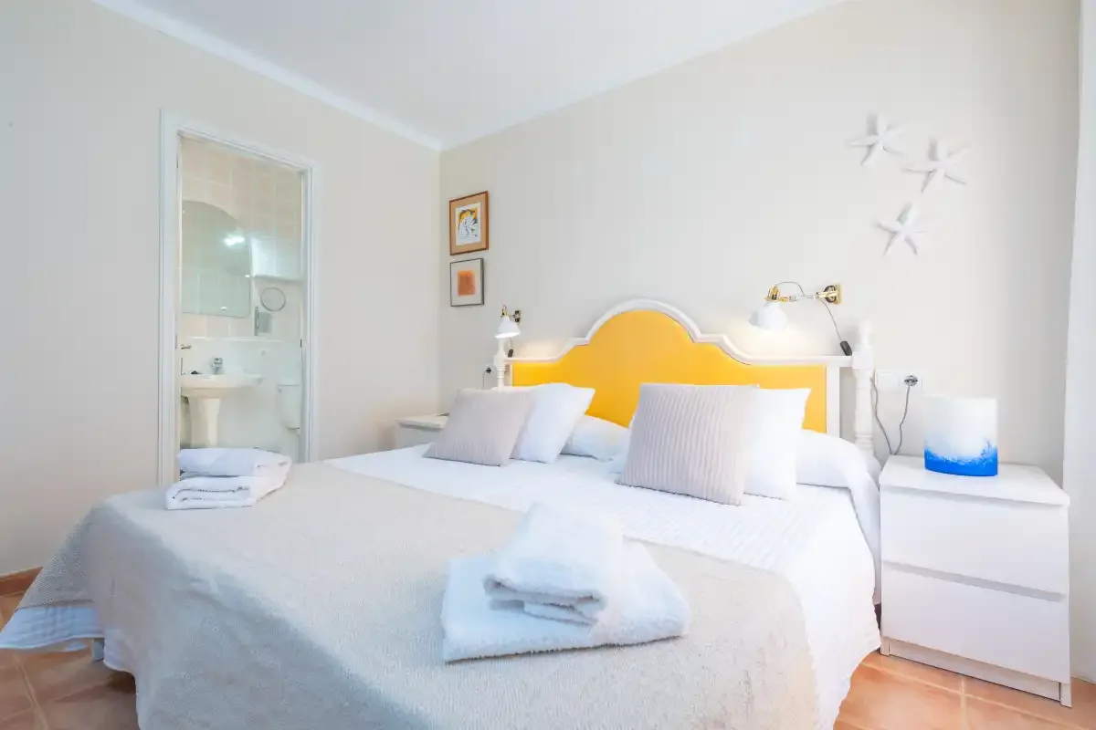 Un dormitorio con una cama blanca, un cabecero amarillo y una puerta abierta al baño.