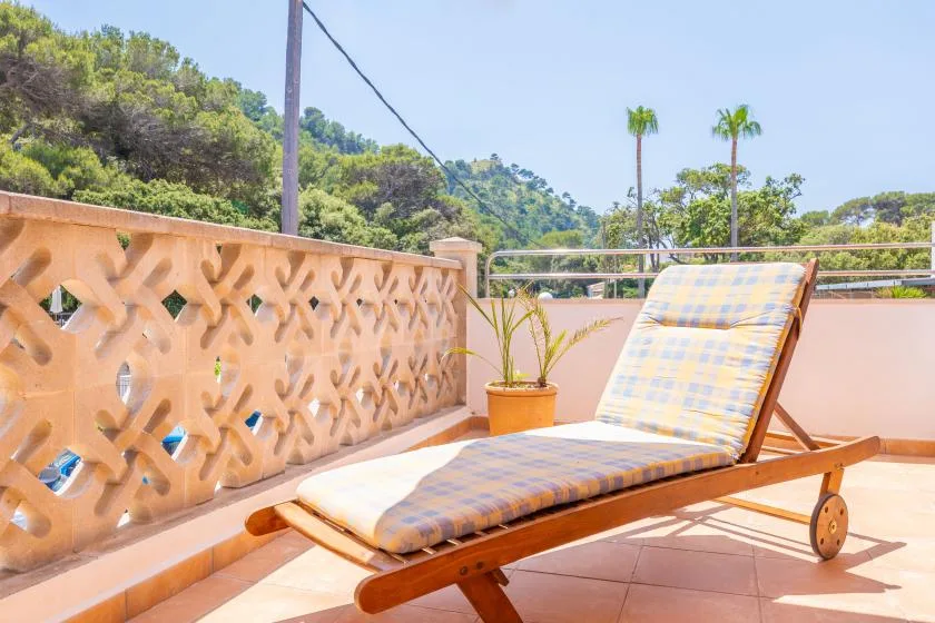 Una terraza embaldosada tiene una tumbona de madera acolchada junto a una barandilla decorativa.